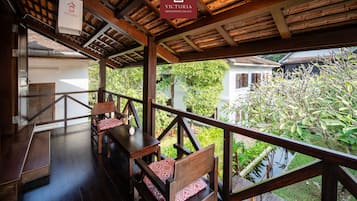 An outdoor wooden deck with a view of a garden and a building. There are wooden benches and a table with a cushion.