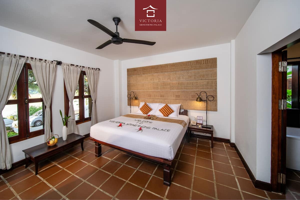 A hotel room with a bed, a ceiling fan, a nightstand, a wall-mounted lamp, and a sign reading "Victoria Xieenthong Palace".