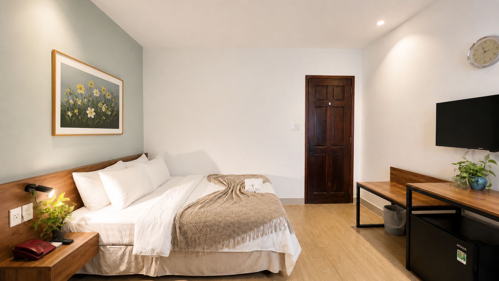 A hotel room with a bed, bedside table, lamp, TV, and a door.