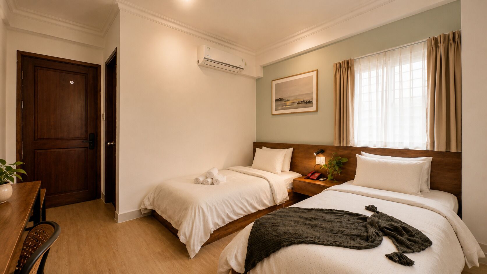 A hotel room with two beds, a wooden door, a small table with a plant, and a painting on the wall.