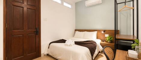 Habitación de hotel con puerta de madera, cama con ropa blanca, mesita de noche con lámpara, una planta pequeña y una lámpara colgante.