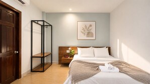 Quarto de hotel com porta de madeira, mesa de cabeceira com abajur, cama com roupa de cama branca e uma obra de arte emoldurada na parede.