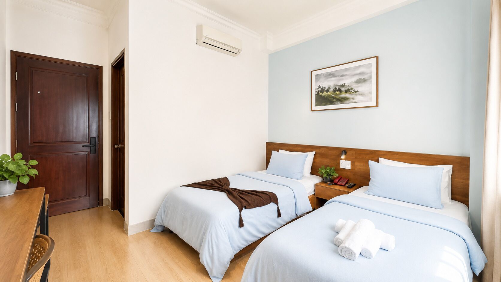 A hotel room with two beds, a wooden door, a small table with a plant, and a painting on the wall.
