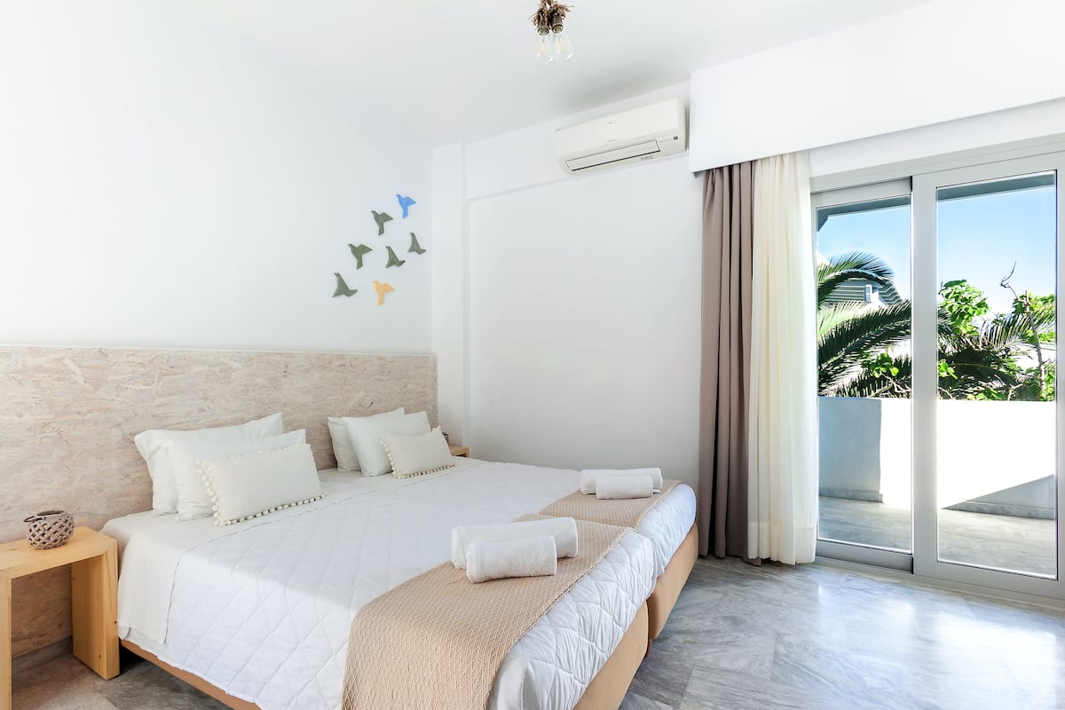 A modern hotel room with a large bed, a wooden nightstand, a wall-mounted air conditioner, and a view of palm trees through the balcony doors.