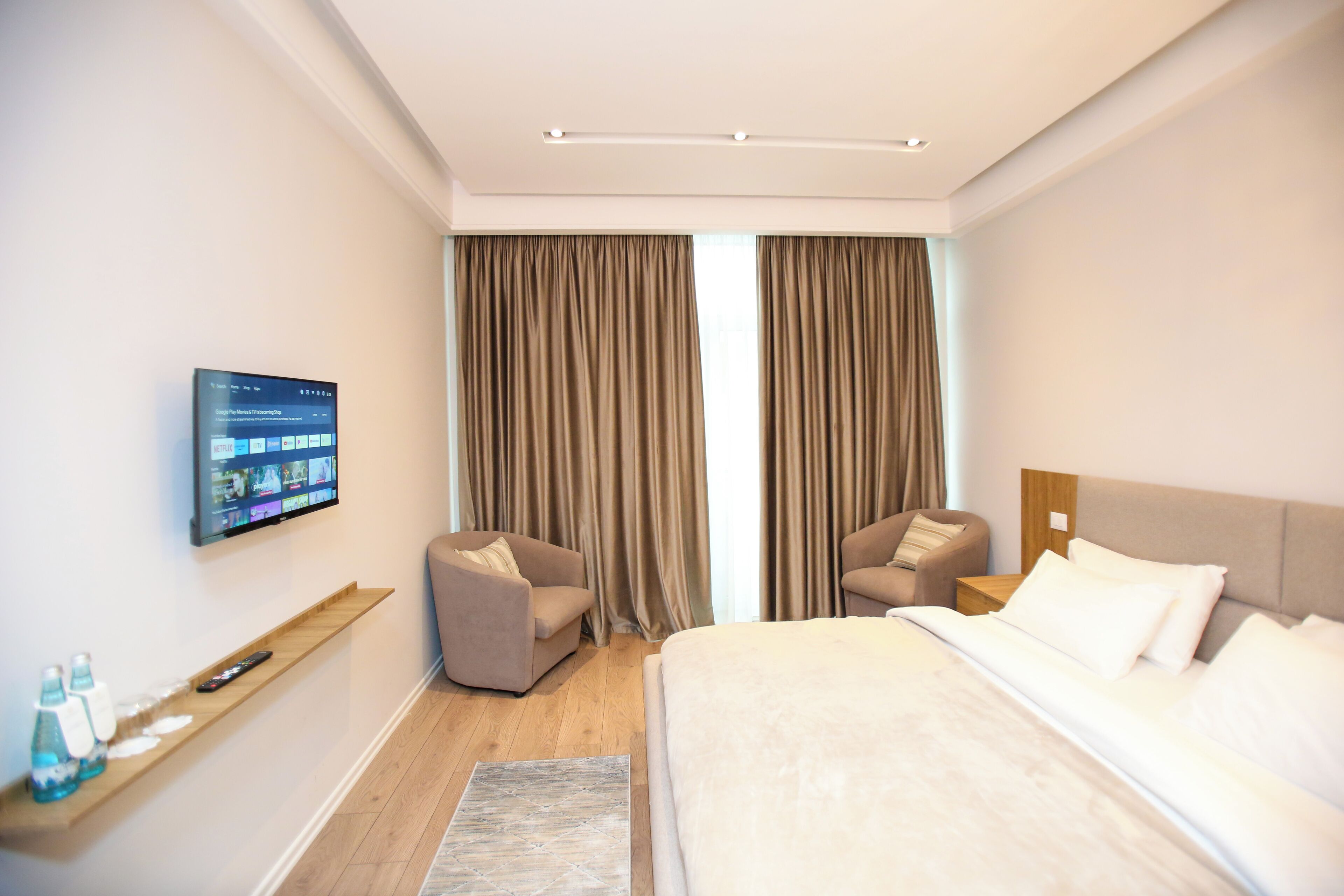 A modern hotel room with a large bed, two armchairs, a flat-screen TV mounted on the wall, and a wooden shelf with a remote control and water bottles.