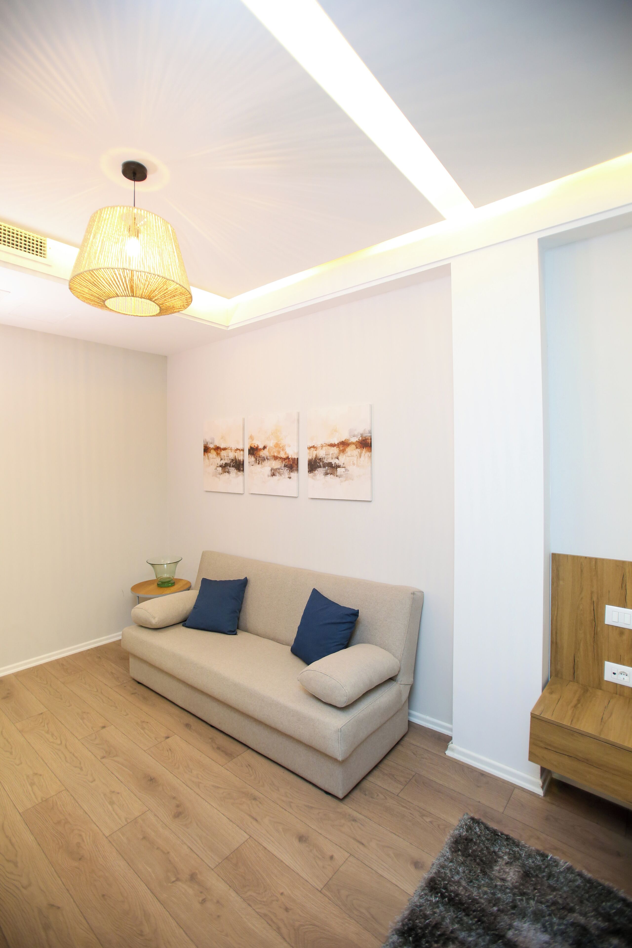 A modern living room with a beige sofa, blue cushions, a glass side table, and a hanging light fixture.