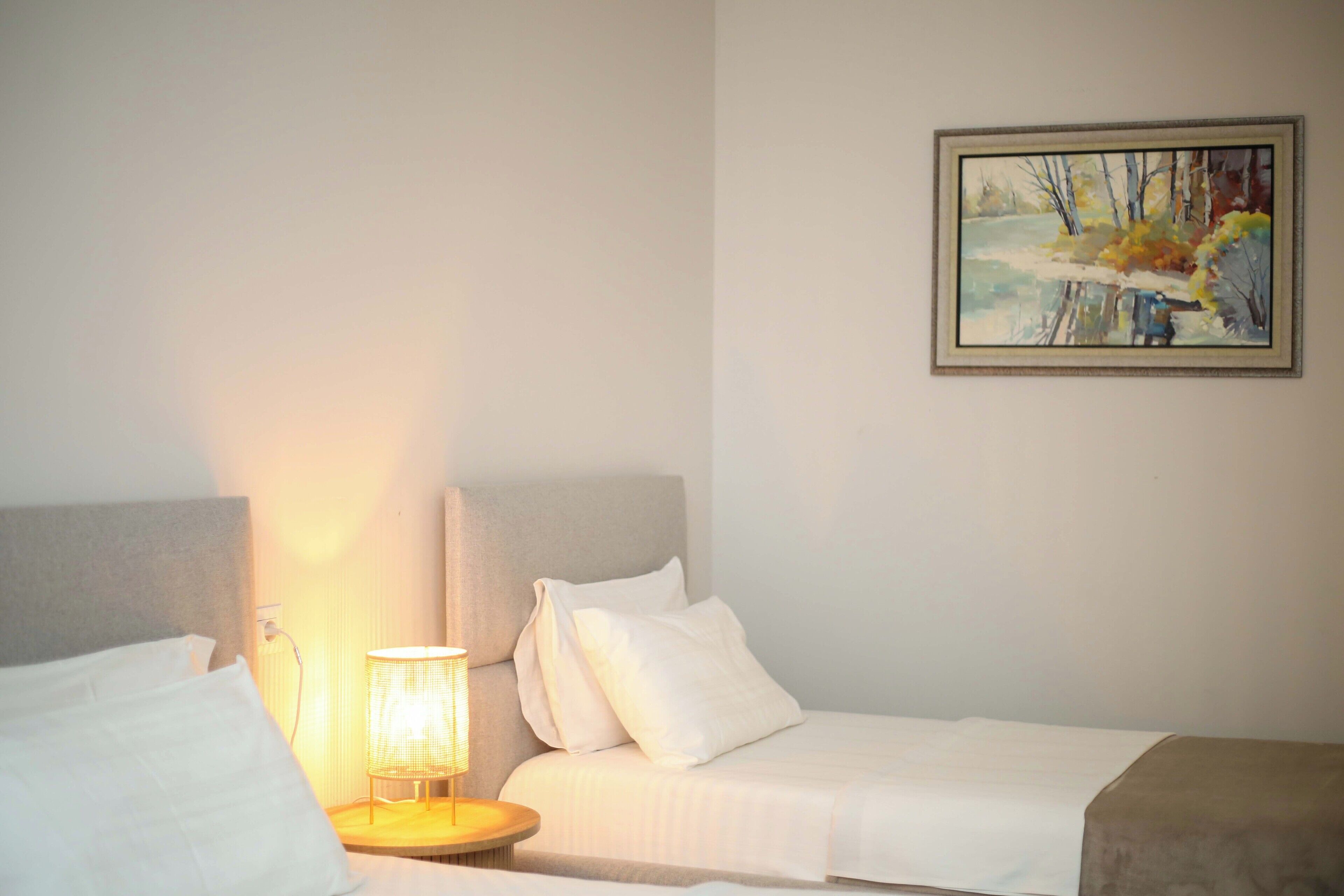 A hotel room with two beds, a bedside table with a lamp, and a painting on the wall.