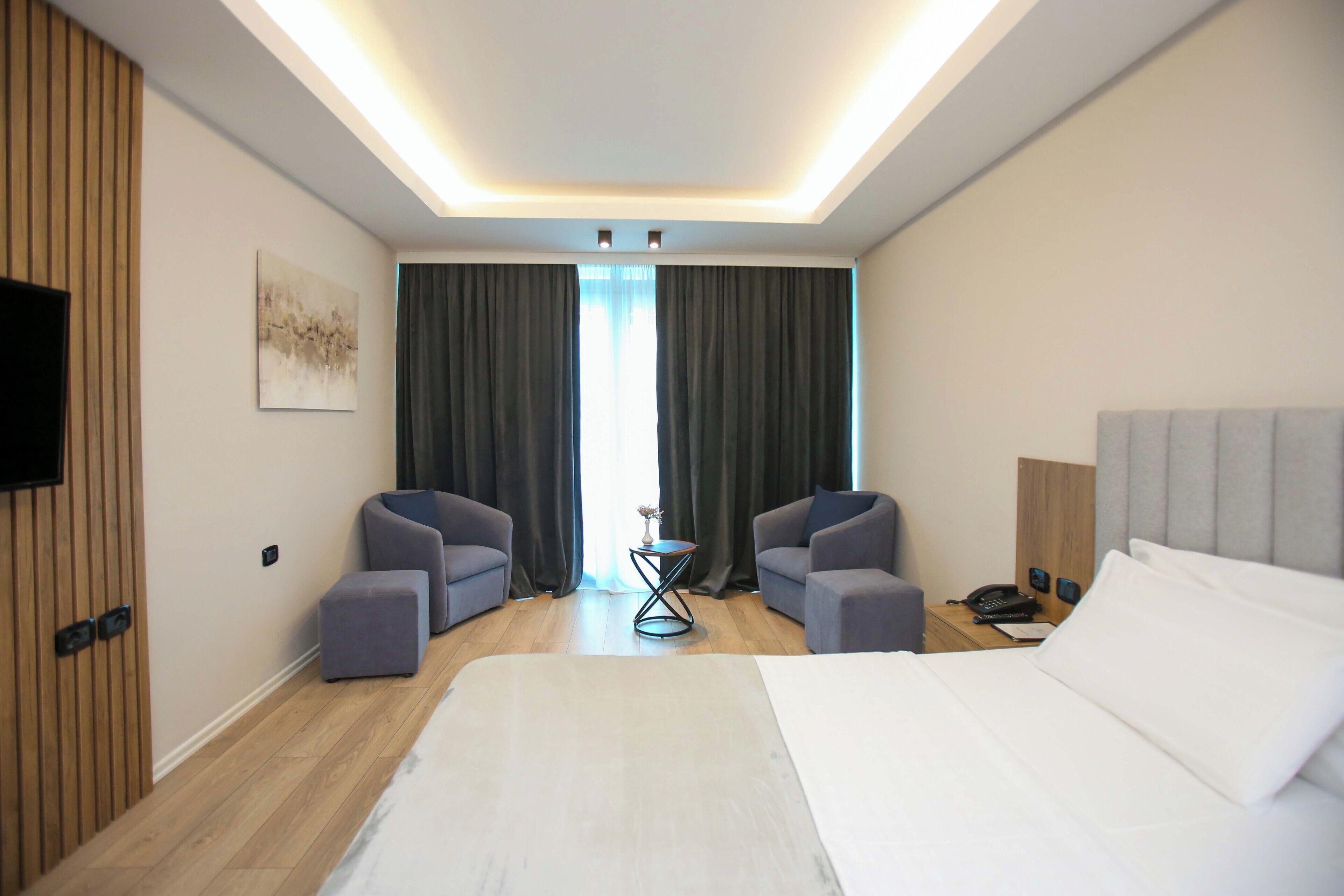 Ein modernes Hotelzimmer mit einem großen Bett, zwei Sesseln, einem kleinen Beistelltisch, einem Flachbildfernseher an der Wand und einem großen Fenster mit Vorhängen.