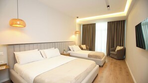 A hotel room with a large bed, a smaller bed, a sofa, and a chair. There are two pendant lights and a large window with curtains. - Vlora International  (Vlorë)