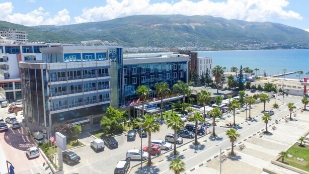 A modern building with a glass facade, surrounded by a parking lot with palm trees, and a view of the sea and mountains in the background. - Vlora International  (Vlorë)