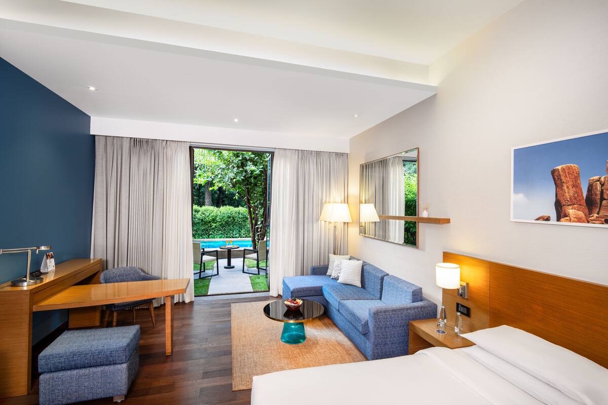 A modern hotel room with a bed, a sofa, a desk, and a dining area with a pool view.