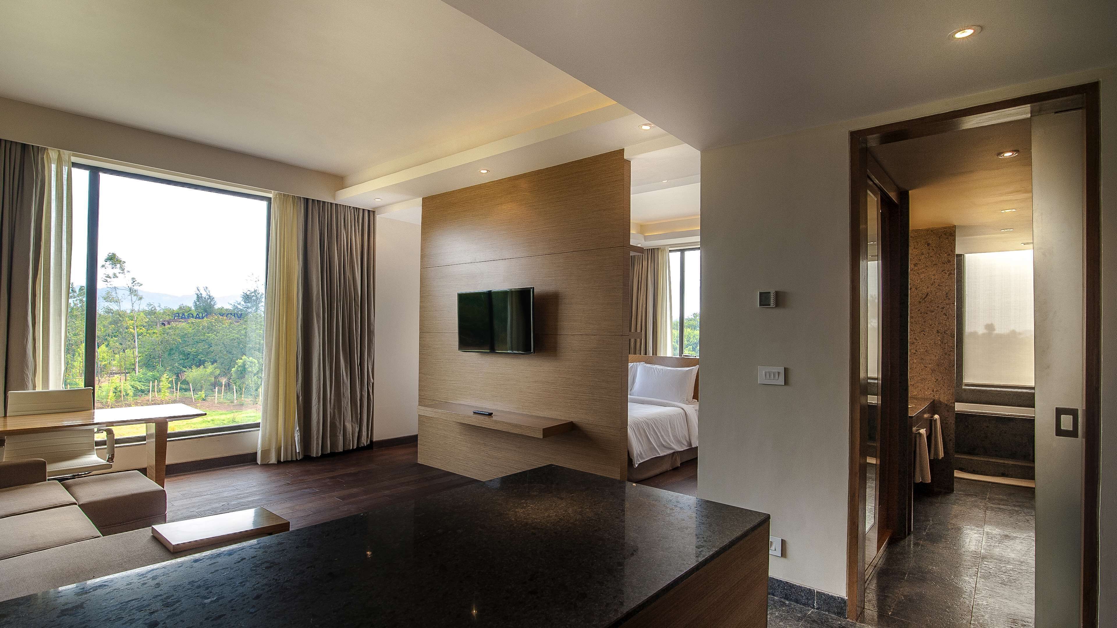 A modern hotel room with a large bed, a flat-screen TV mounted on a wooden panel, a dining area with a table and chairs, and a view of the outdoors through large windows.