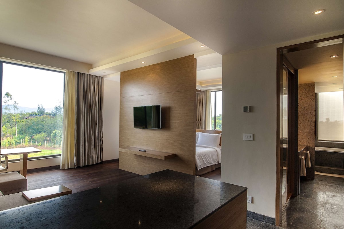 Ein modernes Hotelzimmer mit einem großen Bett, einem an einer Holzvertäfelung befestigten Flachbildfernseher, einem Essbereich mit Tisch und Stühlen sowie einem Blick ins Freie durch große Fenster.