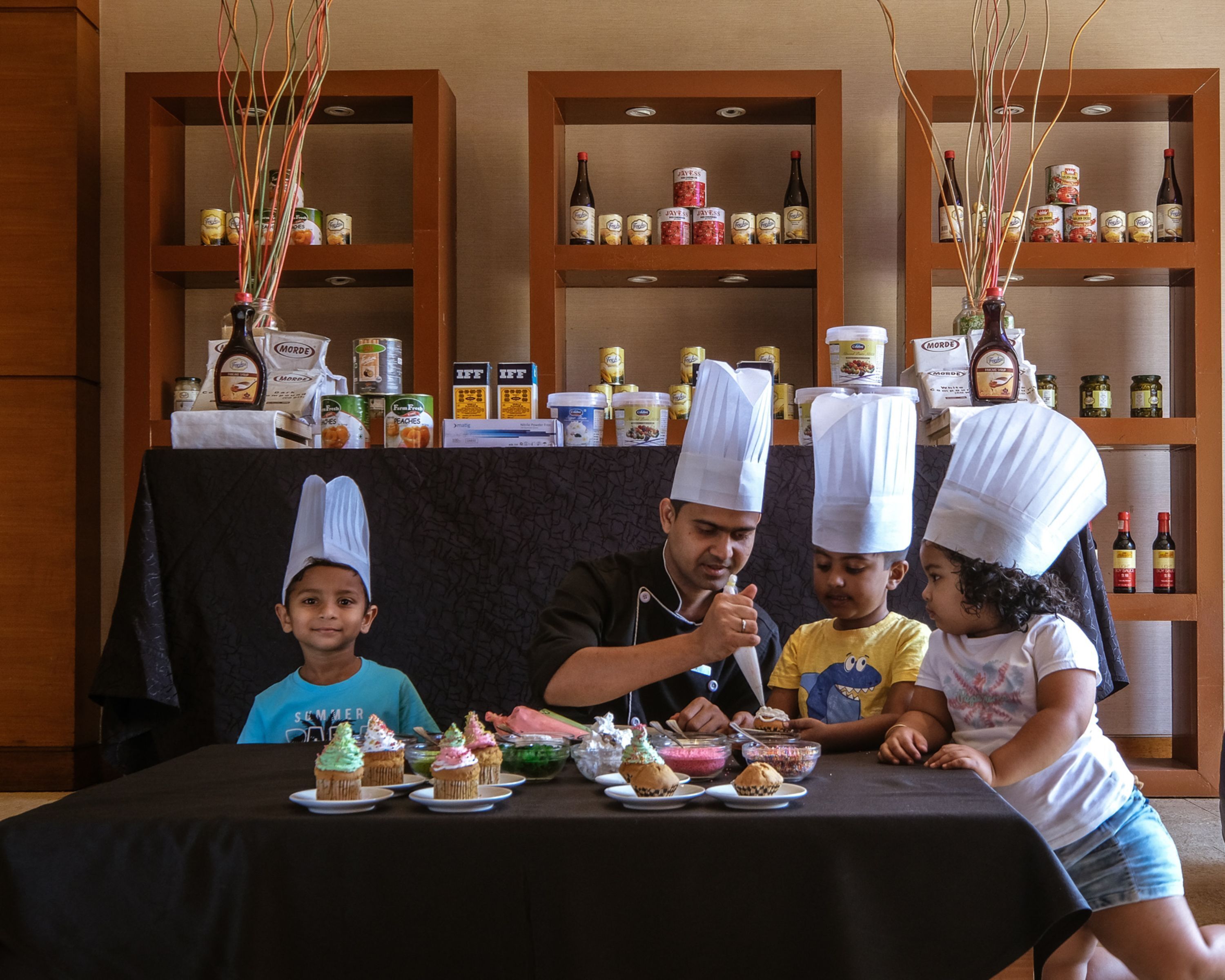Un grupo de personas sentadas a una mesa con diferentes alimentos y bebidas, con estantes al fondo llenos de botellas y latas.