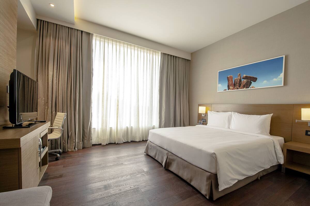 A hotel room with a large bed, a desk with a chair, a television, and a painting on the wall.