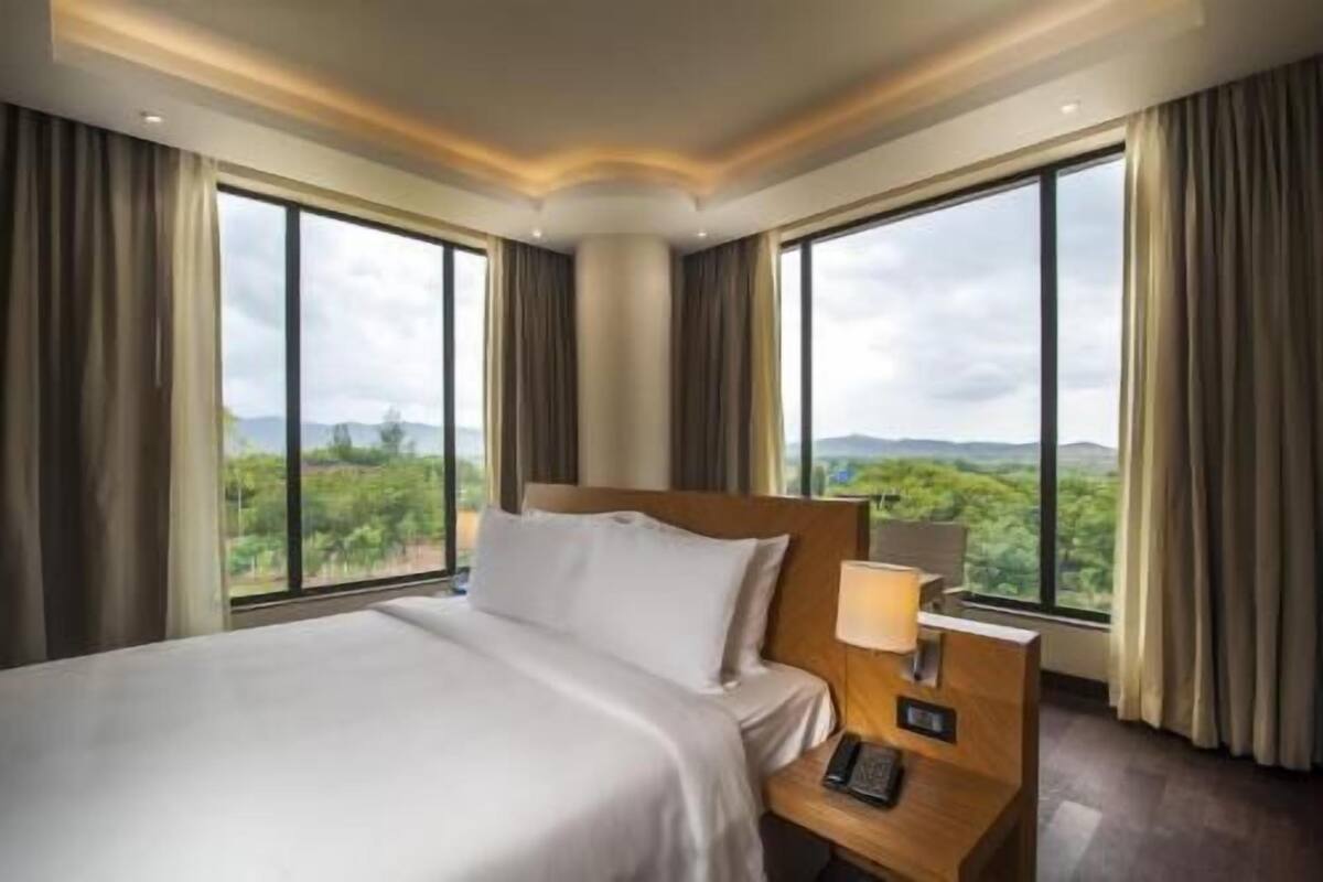 A hotel room with a large bed, a bedside table with a lamp, and a view of greenery through large windows.