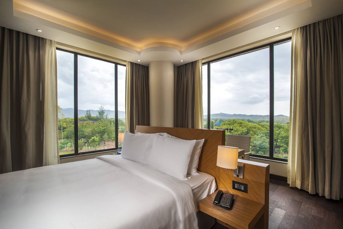 A hotel room with a large bed, a bedside table with a lamp, and a view of the mountains through large windows.