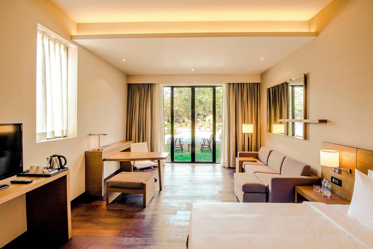 A hotel room with a bed, a desk, a chair, a sofa, a TV, and a balcony with curtains.