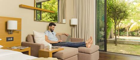A hotel room with a bed, a sofa, a desk with a laptop, and a large window with a view of greenery.
