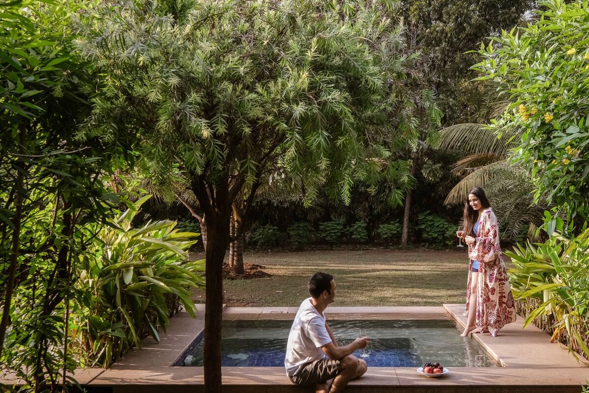 A man sitting by a pool with a woman standing nearby, surrounded by lush greenery.