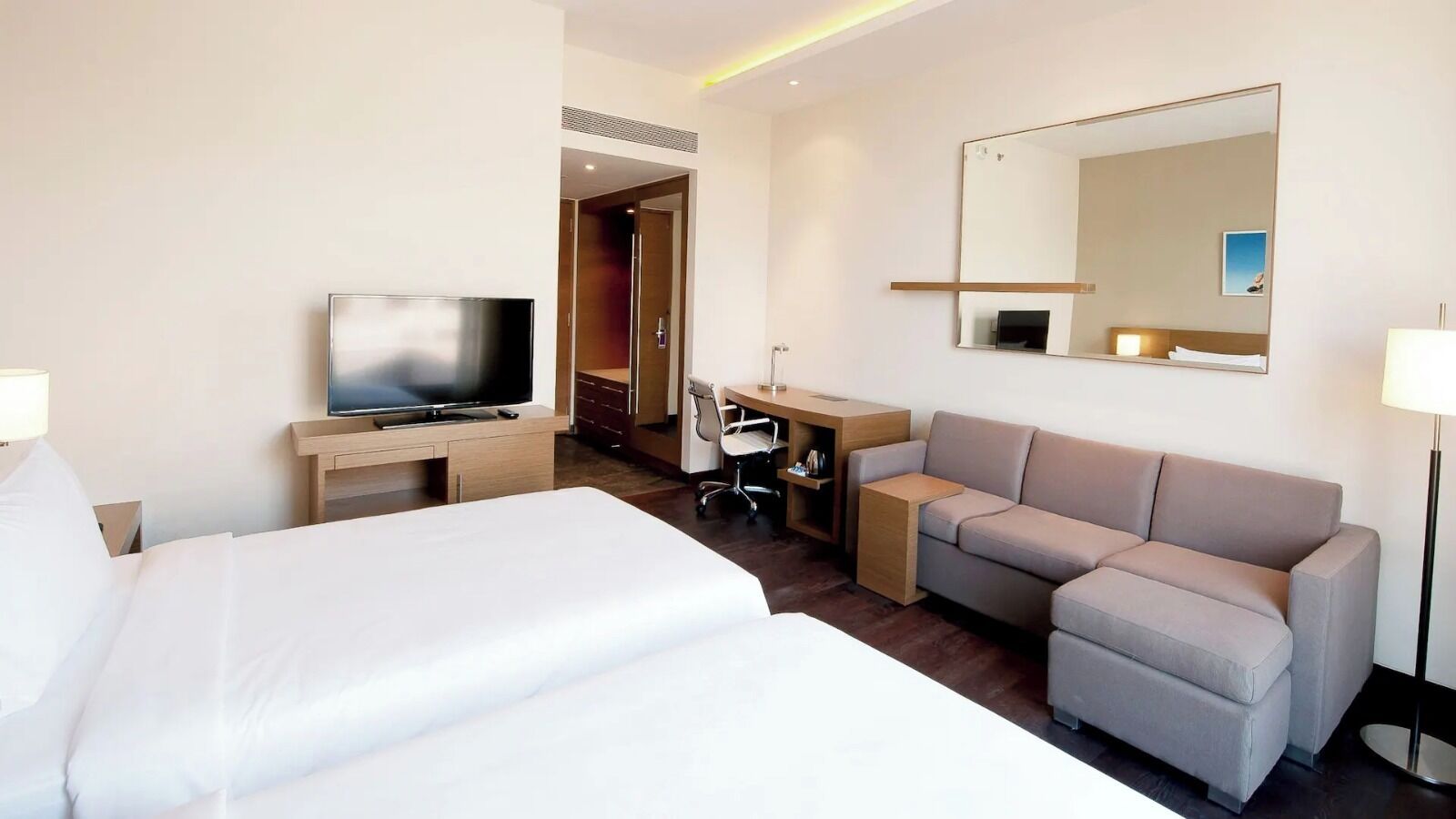 A hotel room with a large bed, a desk with a computer, a sofa, a TV, and a mirror.