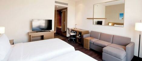 A hotel room with a large bed, a desk with a computer, a sofa, a TV, and a mirror.
