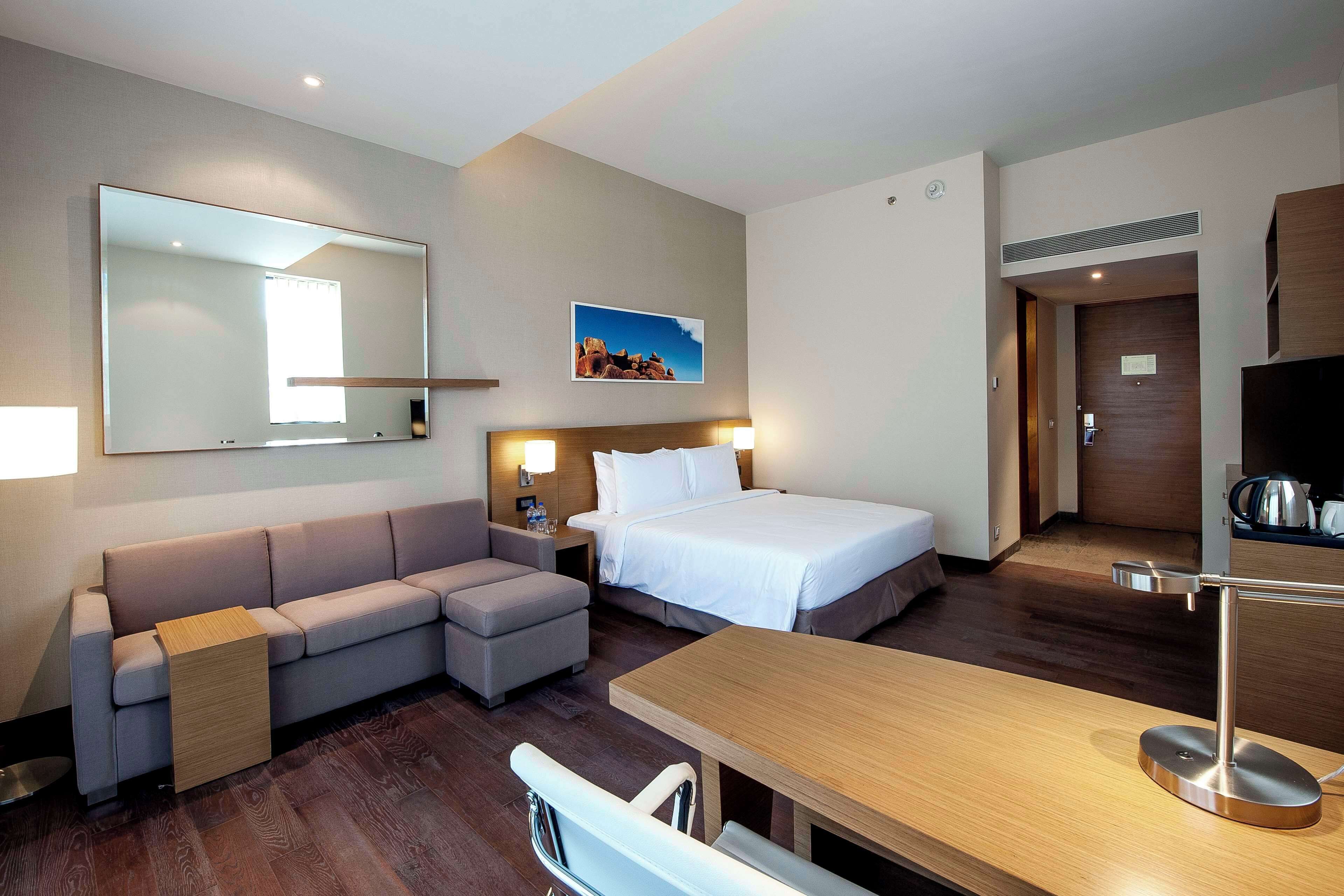 A modern hotel room with a large bed, a sofa, a desk, and a mirror.
