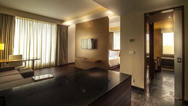 A modern hotel room with a large dark marble countertop, a flat-screen TV mounted on a wooden panel, a bed with white linens, and a view of the hallway. - Hyatt Place Hampi (Sandur)