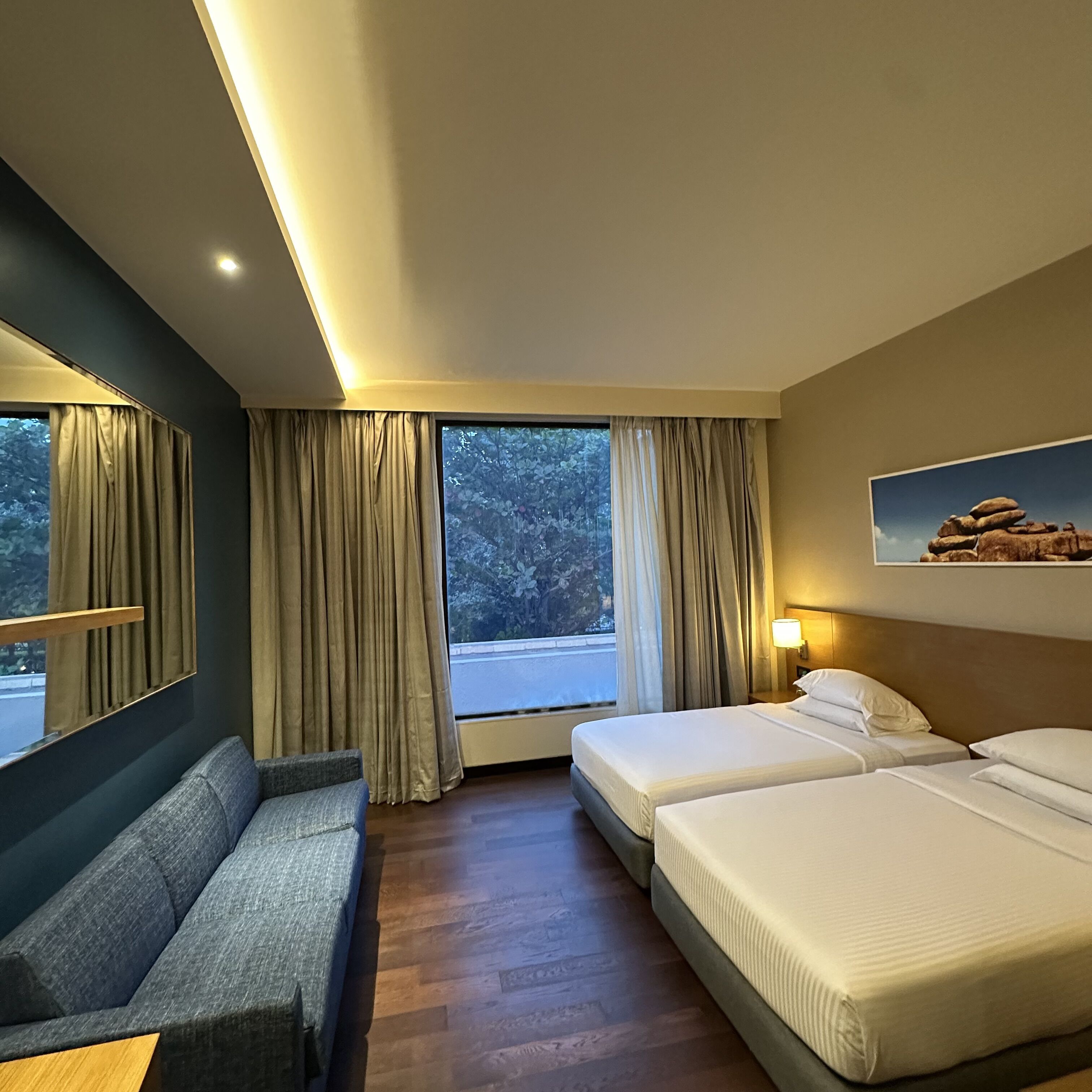 Ein Hotelzimmer mit einer Couch, zwei Betten, einem großen Fenster mit Vorhängen, einer Wandlampe und einem gerahmten Bild.