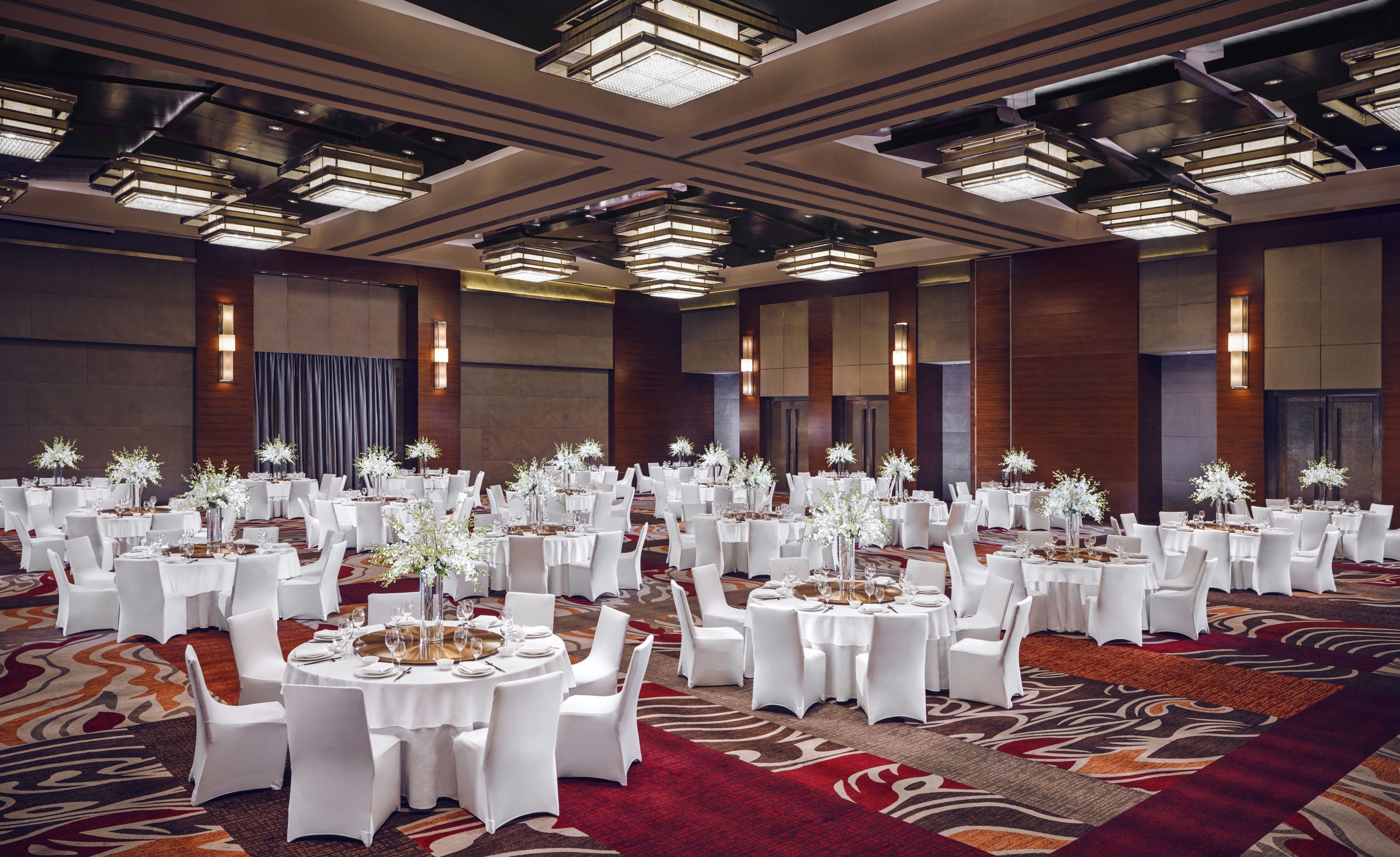 Une grande salle de banquet avec des tables rondes, des chaises blanches et des centres de table ornés de compositions florales.
