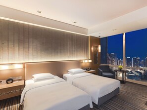 A hotel room with two beds, a desk, and a chair. There is a city view through large windows.