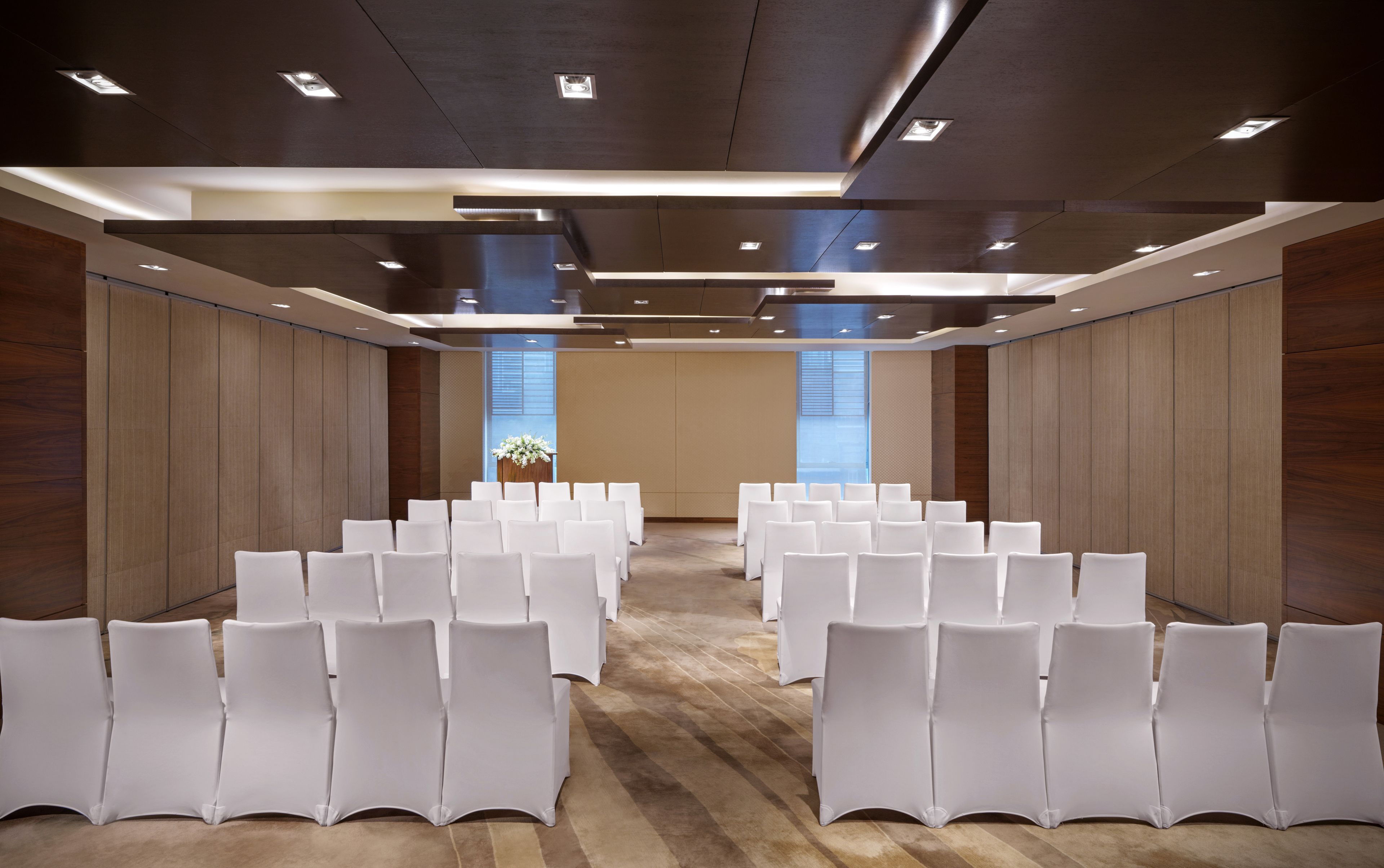 Une salle de conférence avec des rangées de chaises blanches, des murs recouverts de panneaux de bois et un éclairage encastré.
