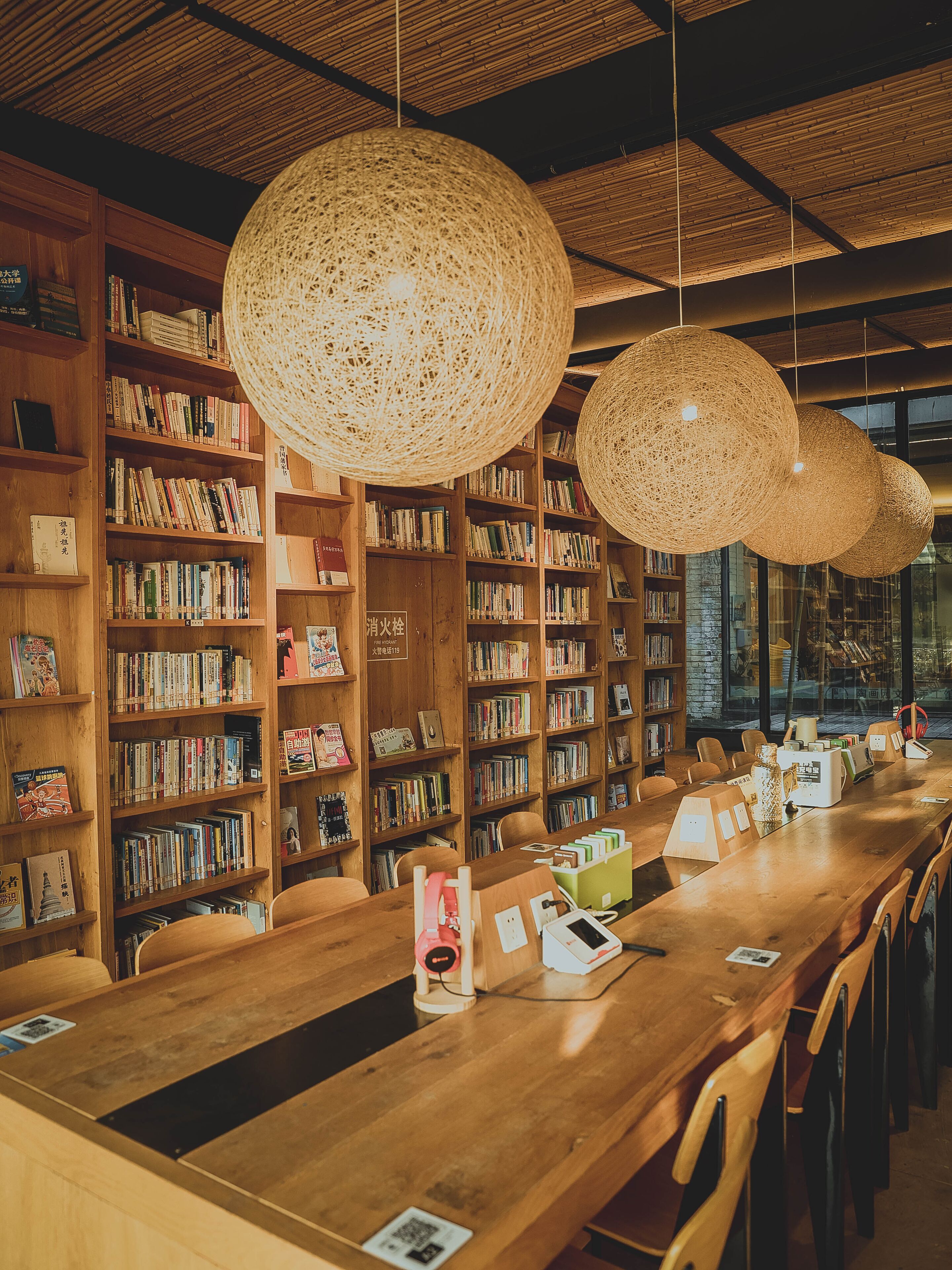 Une bibliothèque avec des étagères en bois, des luminaires suspendus en fibres tressées et une longue table en bois entourée de chaises.