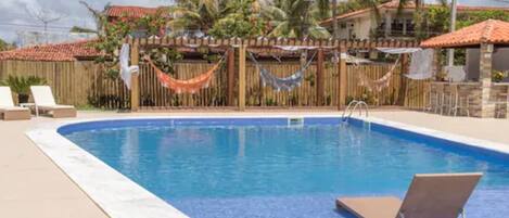 A rectangular swimming pool with a wooden fence and hammock decorations, surrounded by palm trees and a tiled roof building.