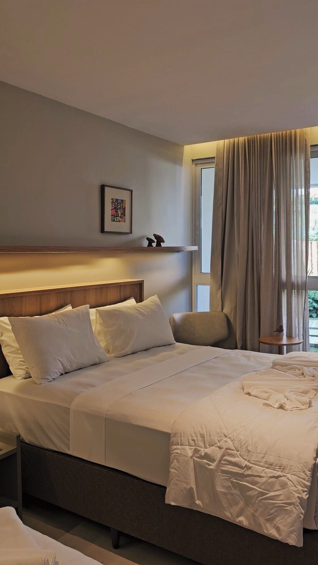 Ein Hotelzimmer mit einem Bett, einem Kopfteil, einem Fenster mit Vorhängen, einem kleinen Tisch und einem an der Wand hängenden Bild.