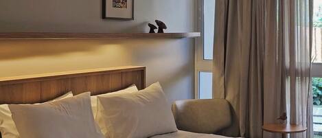 Ein Hotelzimmer mit einem Bett, einem Kopfteil, einem Fenster mit Vorhängen, einem kleinen Tisch und einem an der Wand hängenden Bild.