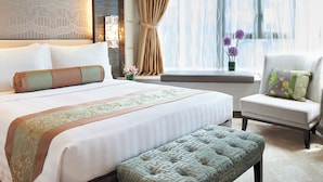 Habitación de hotel con una cama grande, una zona de estar con una silla, un banco, una araña y una ventana con cortinas.