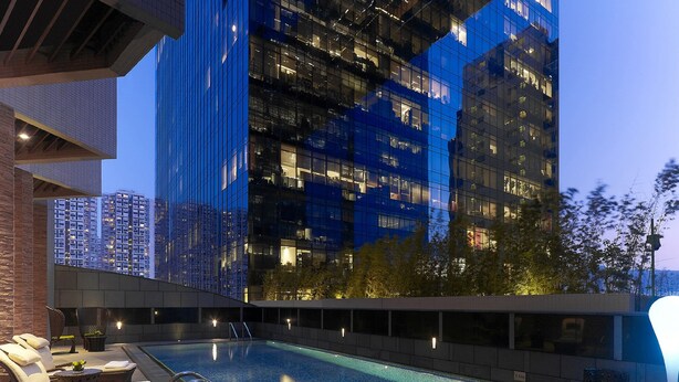 Un área de piscina en la azotea con sillones de descanso y vista a un edificio moderno con ventanales de vidrio reflectante.
