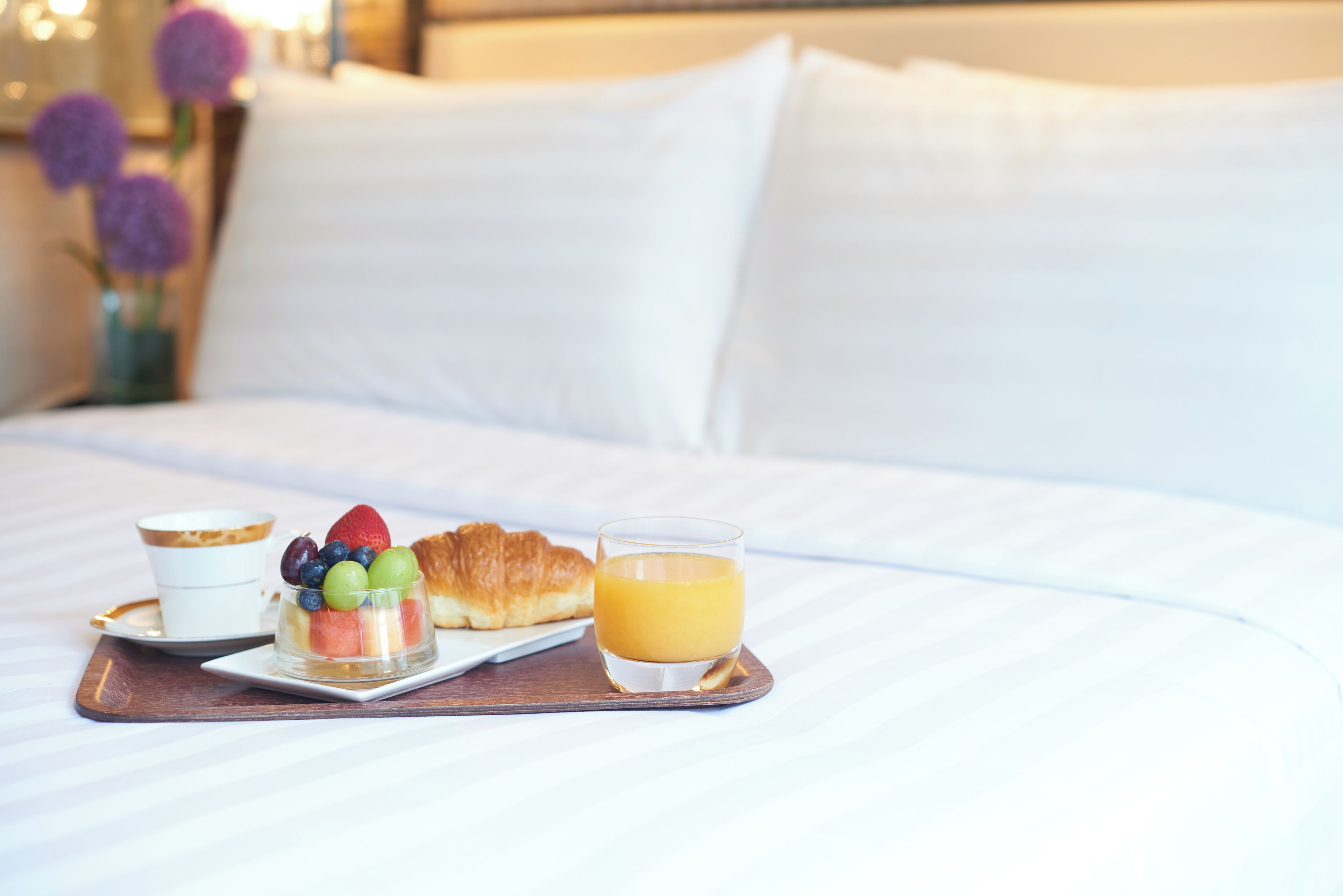 Bandeja de desayuno con un croissant, fruta y jugo de naranja sobre una cama con ropa de cama y almohadas blancas.