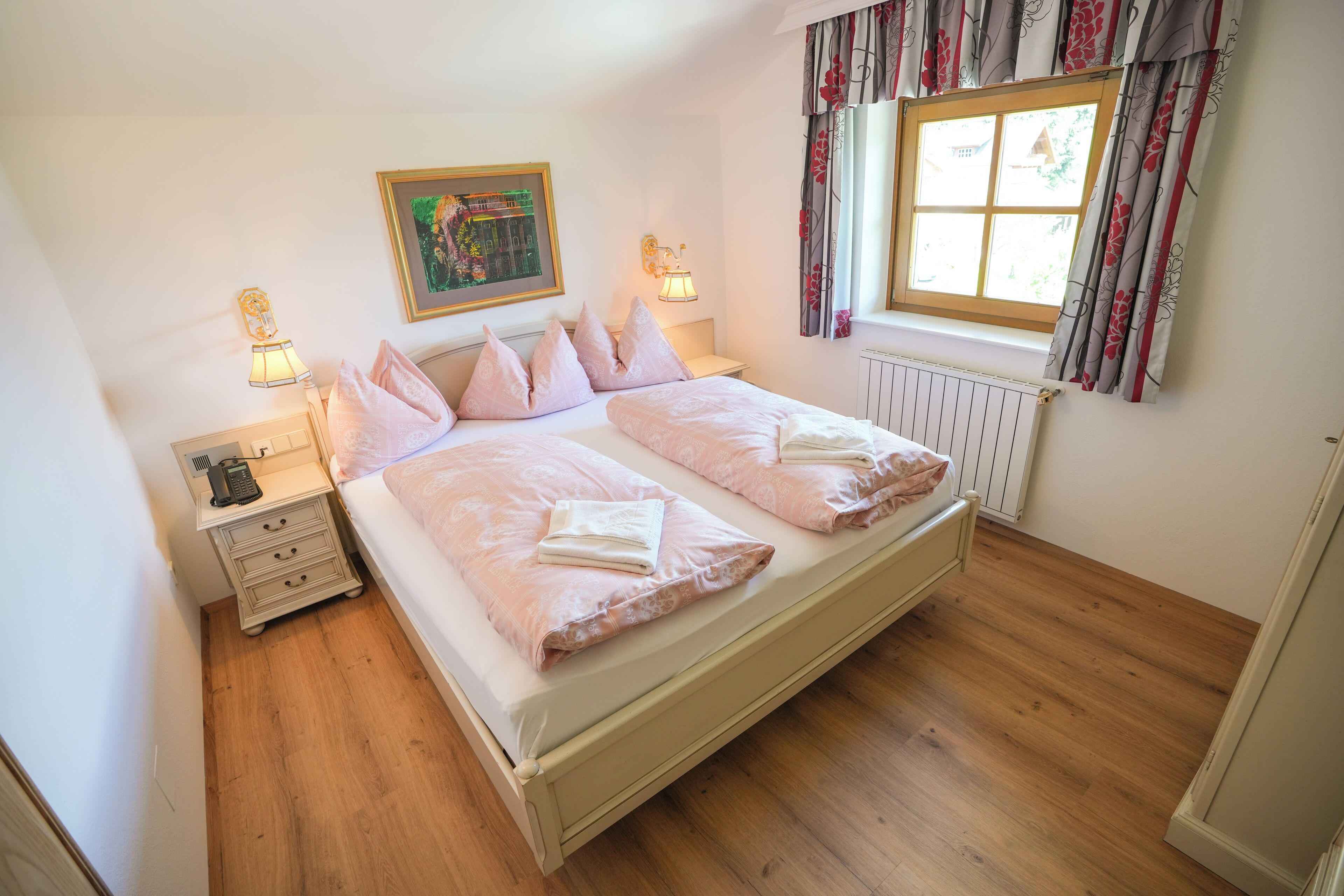 A double bed with a white headboard, two bedside tables with lamps, a framed picture on the wall, and a window with curtains.