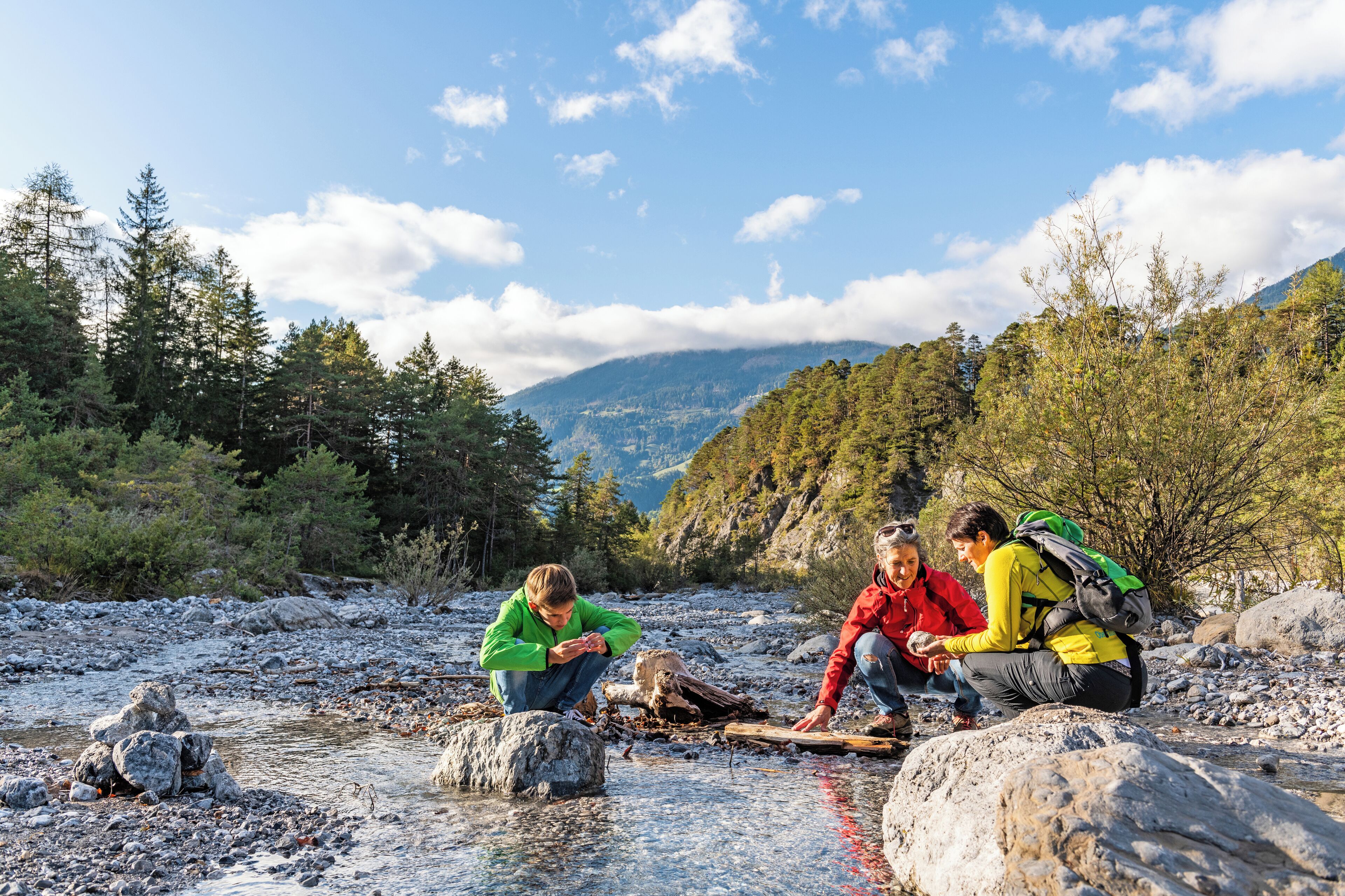 A group of people sitting by a river in a forested area with mountains in the background.