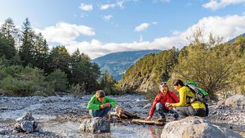 A group of people sitting by a river in a forested area with mountains in the background.