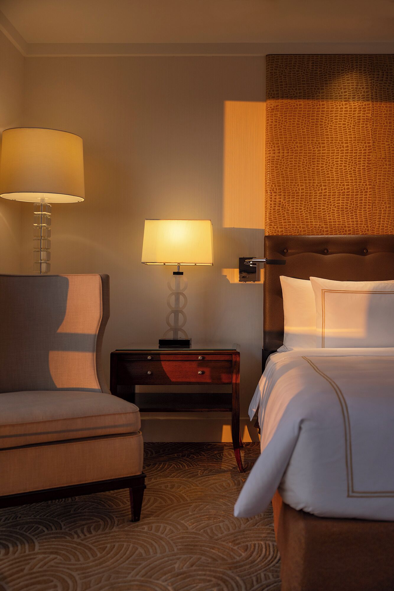A hotel room with a bed, a chair, a nightstand with a lamp, and a wall with a textured pattern.
