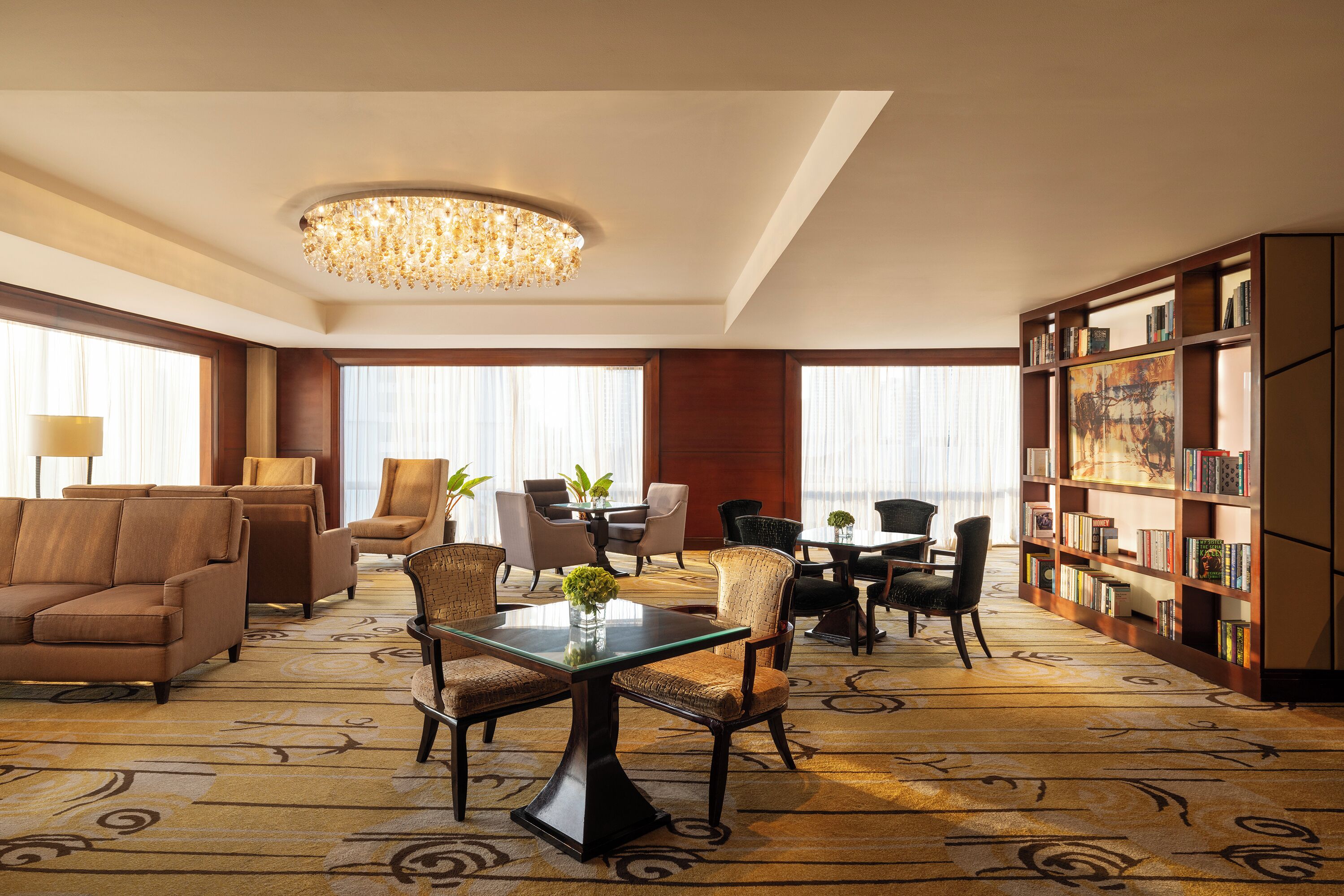 A spacious hotel lobby with a large chandelier, comfortable seating, a central table with a vase, and a bookshelf filled with books.