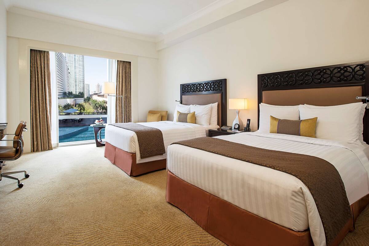 A hotel room with two beds, a desk, and a view of a cityscape through large windows.