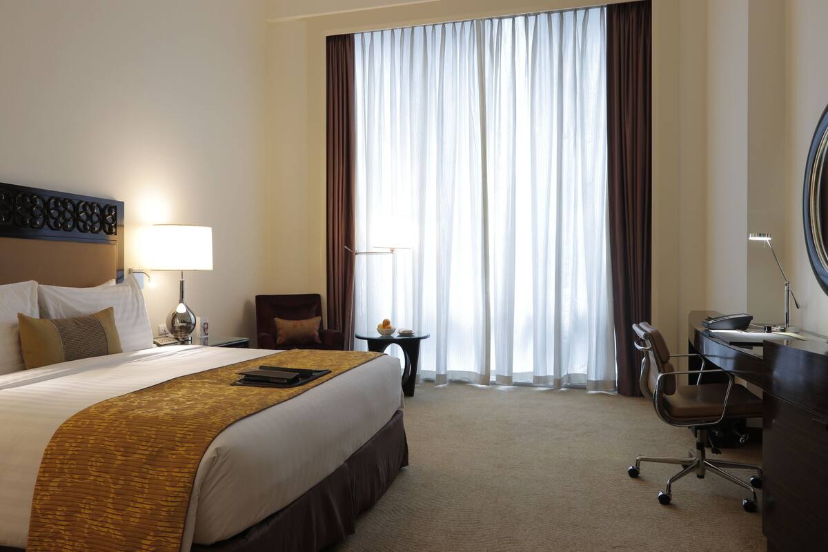 A hotel room with a large bed, a desk, a chair, a lamp, and a view of the outdoors through sheer curtains.