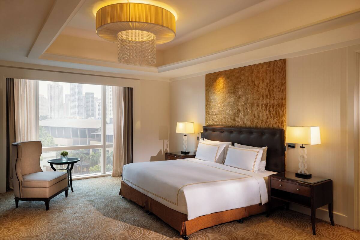 A hotel room with a large bed, two bedside tables with lamps, a chair, a small table with a plant, and a large window with a city view.