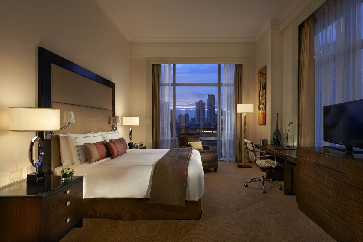 A hotel room with a large bed, two bedside tables, a desk with a chair, a TV, and a city view through large windows.
