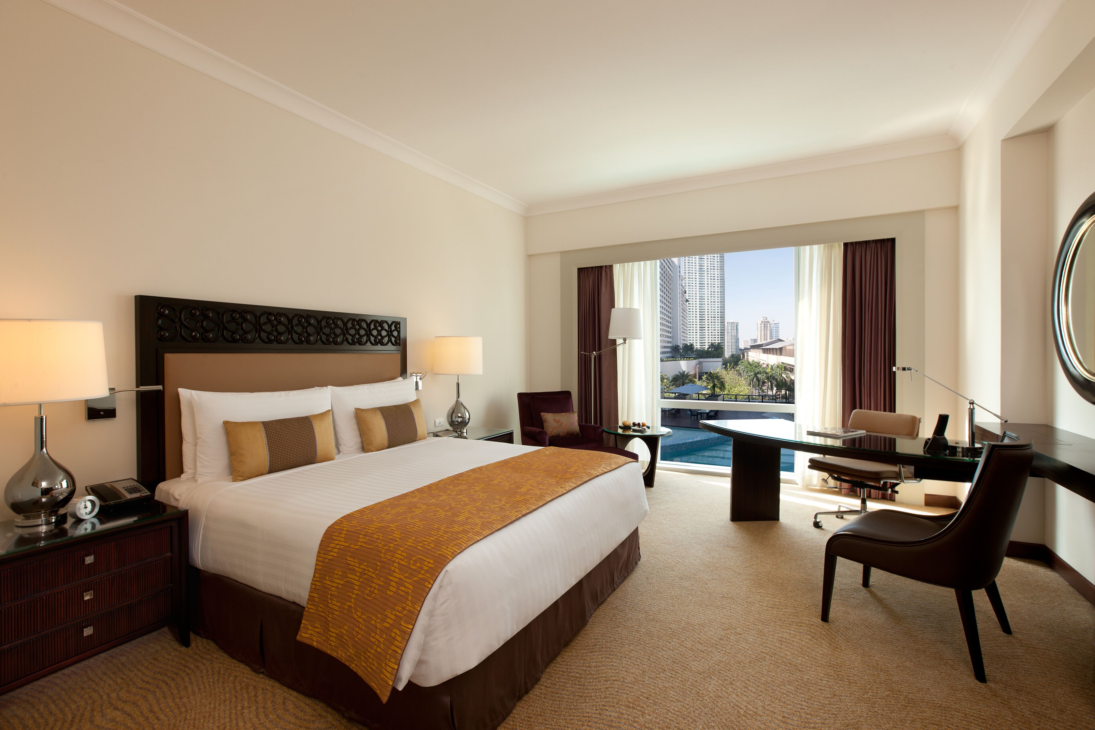 A hotel room with a large bed, a desk with a chair, a view of a cityscape, and a balcony with a pool.