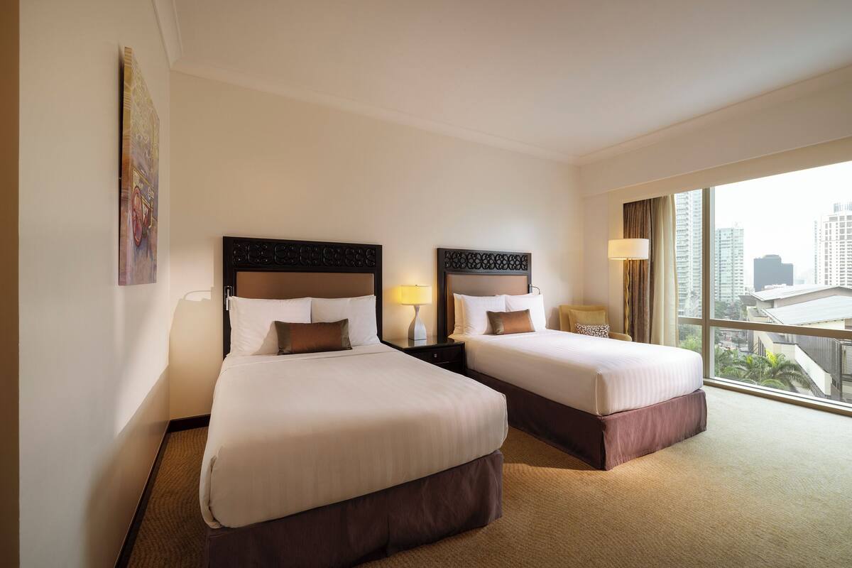 A hotel room with two beds, a large window with a city view, and a painting on the wall.